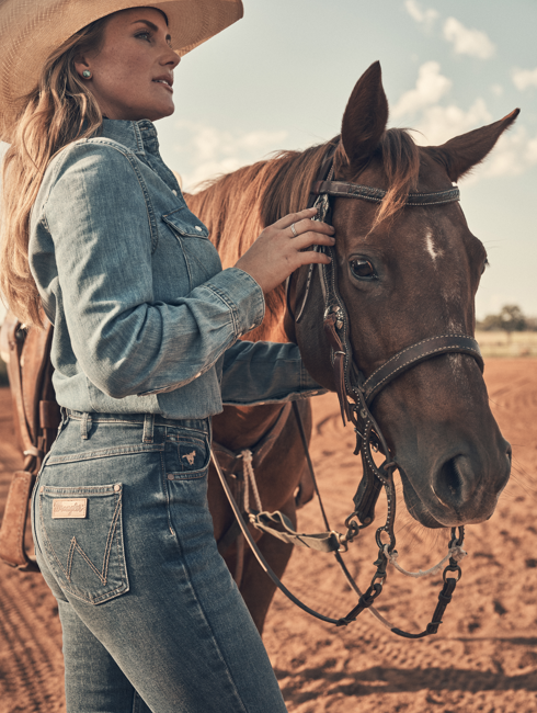 Woman in cowboy hat and leather jacket, seated outdoors, resting on logs.