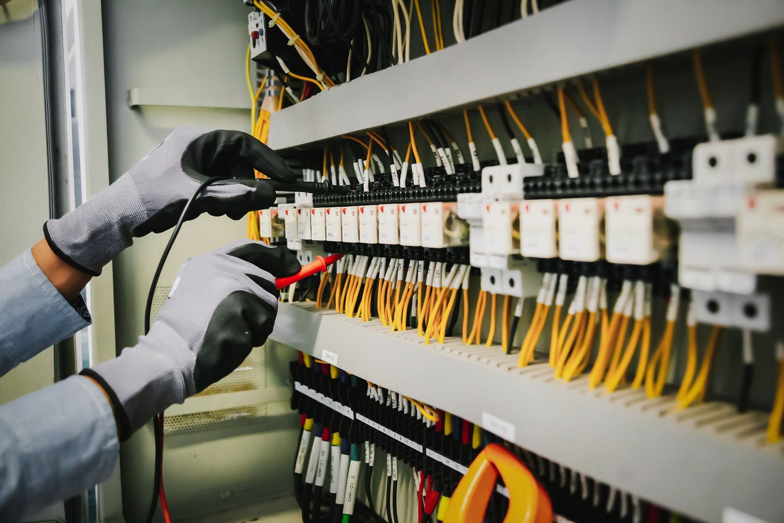 Hands with gloves testing electrical wiring in a control panel.