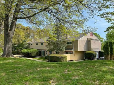 Picture of a beige colored, two-story Heritage Village Landenberg townhouse, surrounded by grass, trees, and hedges on a sunny day.