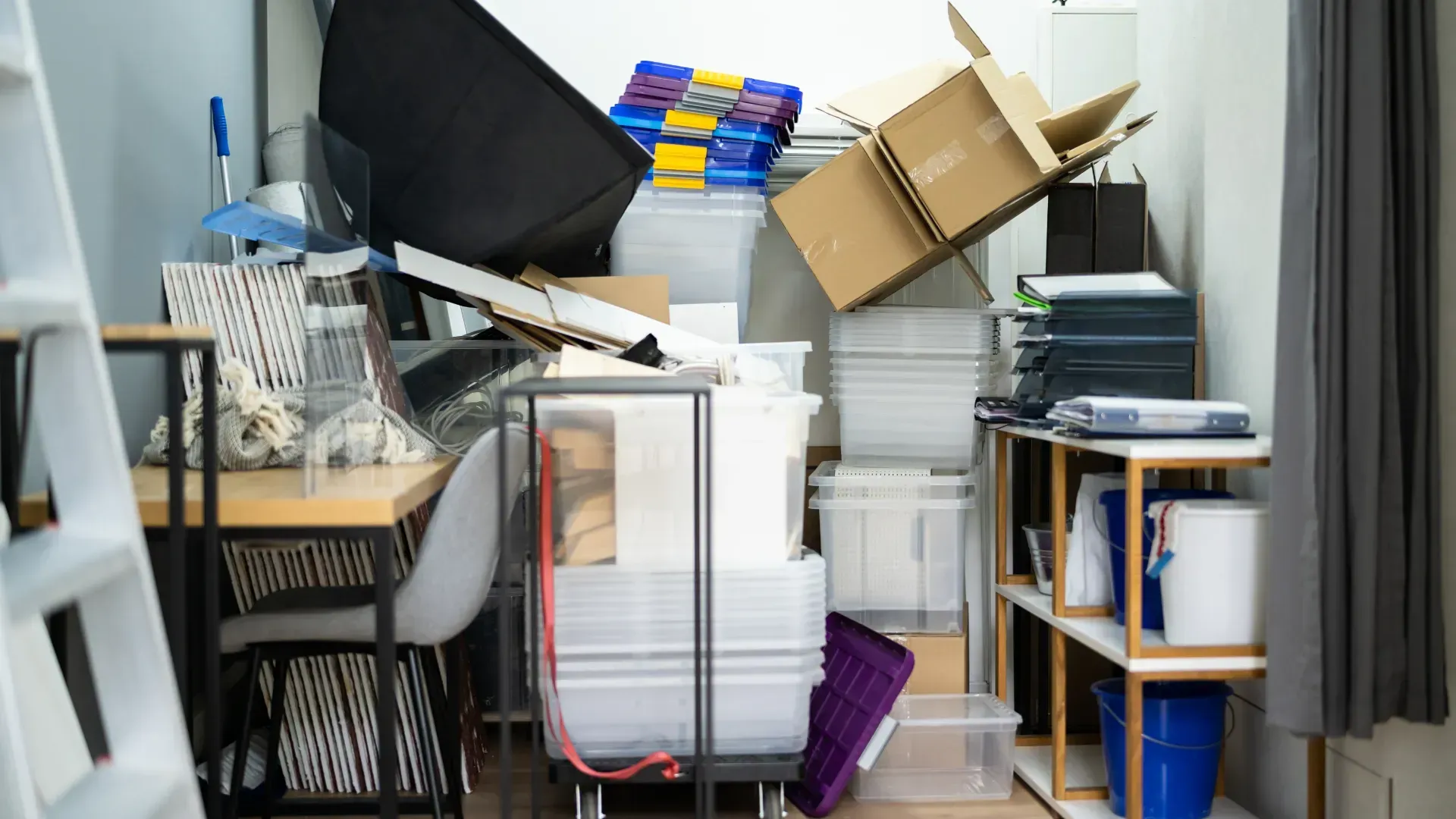 Cluttered room with boxes and items piled high, a desk, and a ladder.