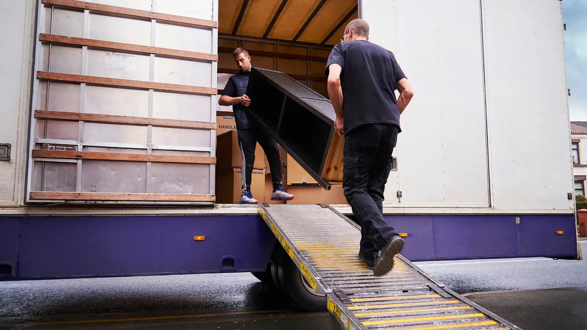 Two men loading a dark cabinet into a moving truck via ramp. Exterior shot.