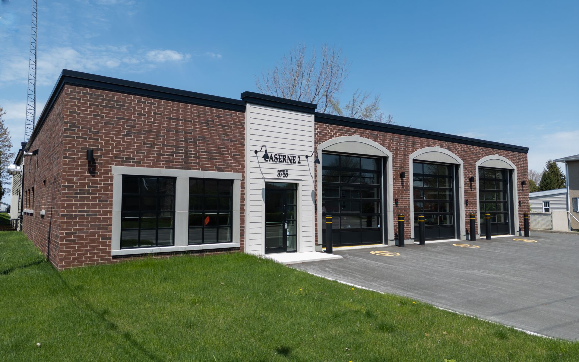 Bâtiment en briques avec trois portes de garage et une porte d'entrée vitrée, pelouse verte et ciel bleu.