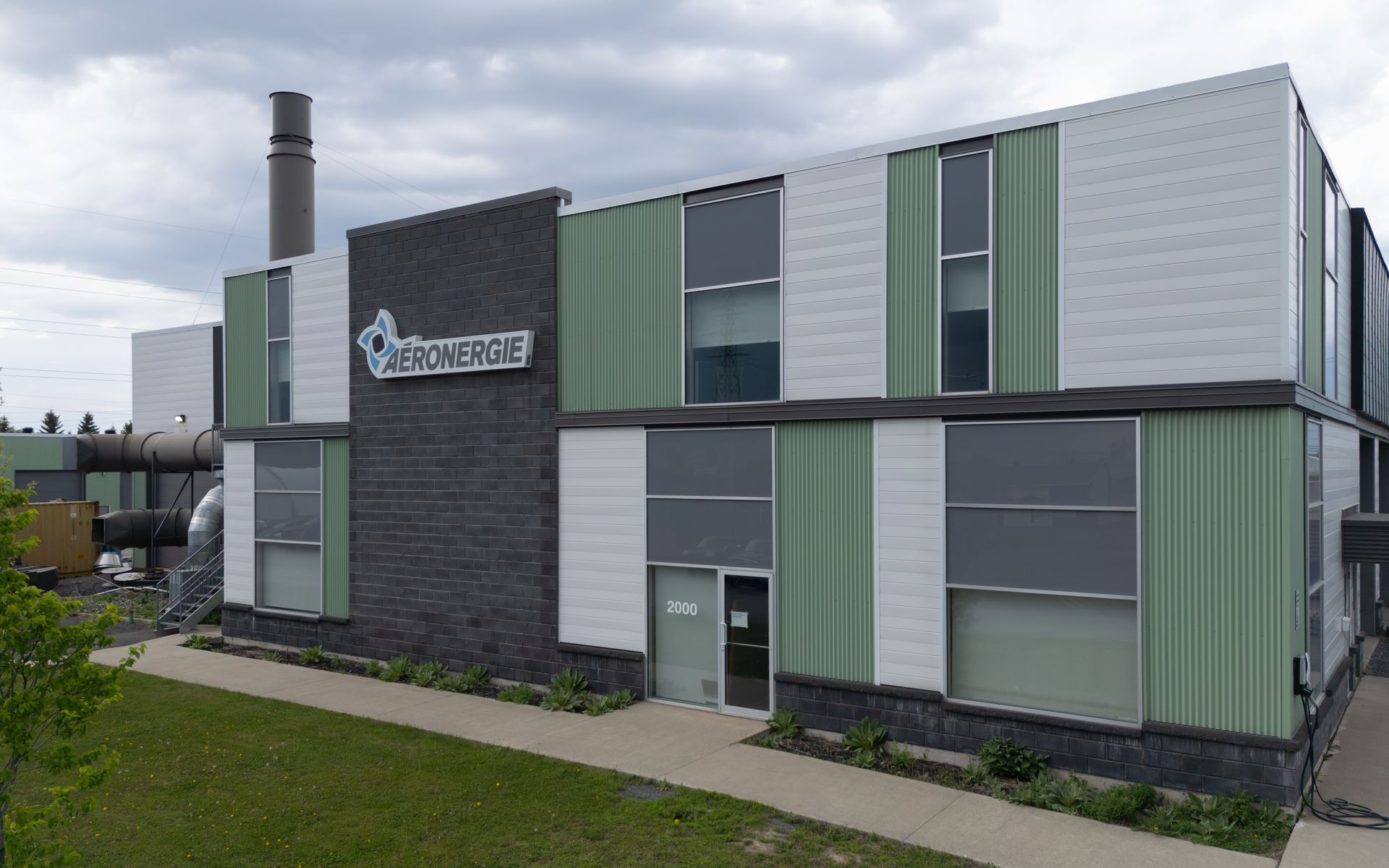 Un bâtiment de deux étages avec des panneaux verts, blancs et gris, une façade en briques et une cheminée. L'enseigne indique « HYDRO ÉNERGIE ».