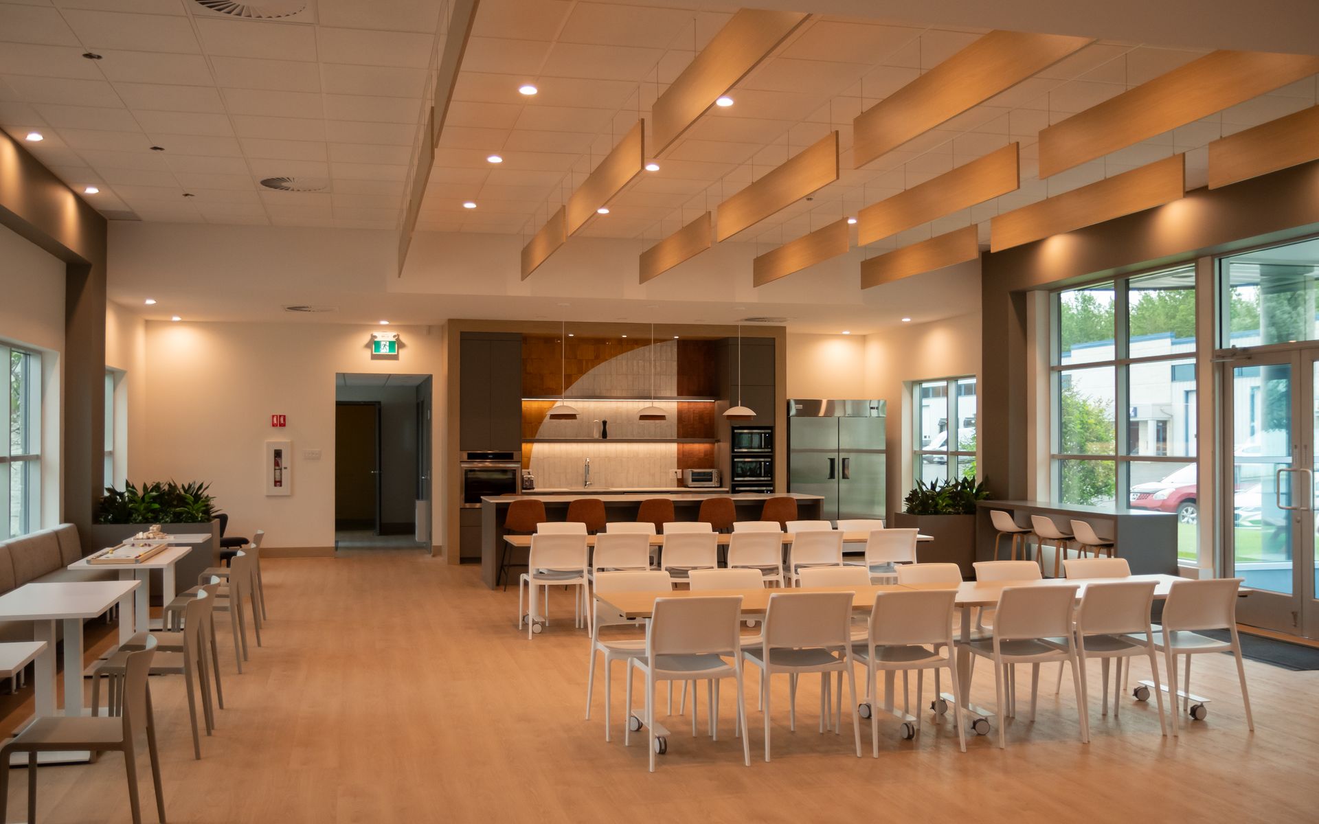 Salle commune vide et bien éclairée, avec tables, chaises, cuisine et grandes fenêtres.