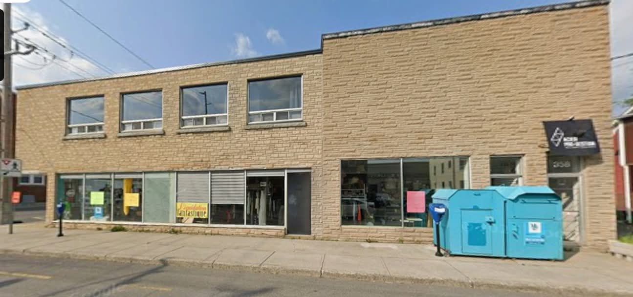 Un bâtiment en briques brun clair avec de grandes fenêtres. Une poubelle de recyclage bleue est placée devant l'une d'elles, à côté d'un panneau noir et blanc.