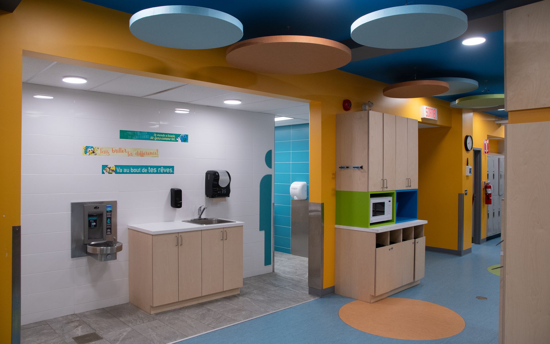 Couloir de garderie aux couleurs vives avec lavabos, armoires et fontaine à eau.