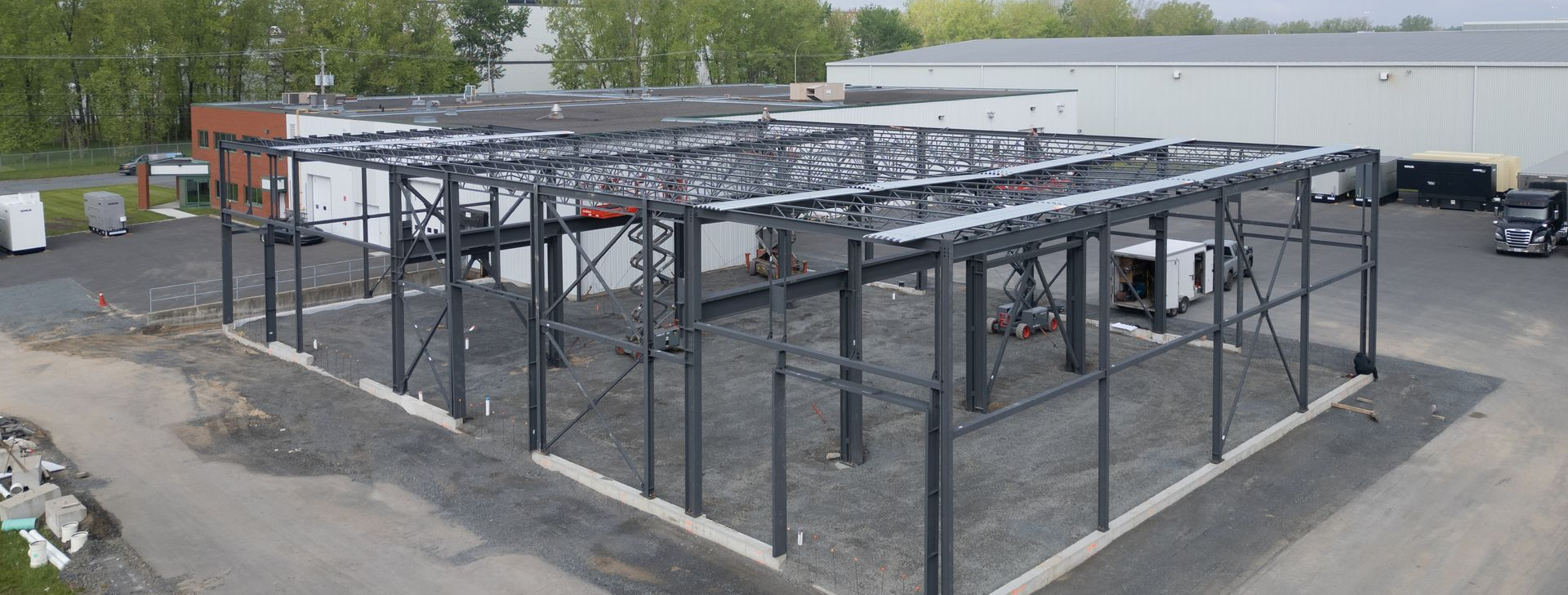 Bâtiment à ossature métallique en construction sur un terrain gravillonné, avec des fermes de toit inachevées et des camions alentour.