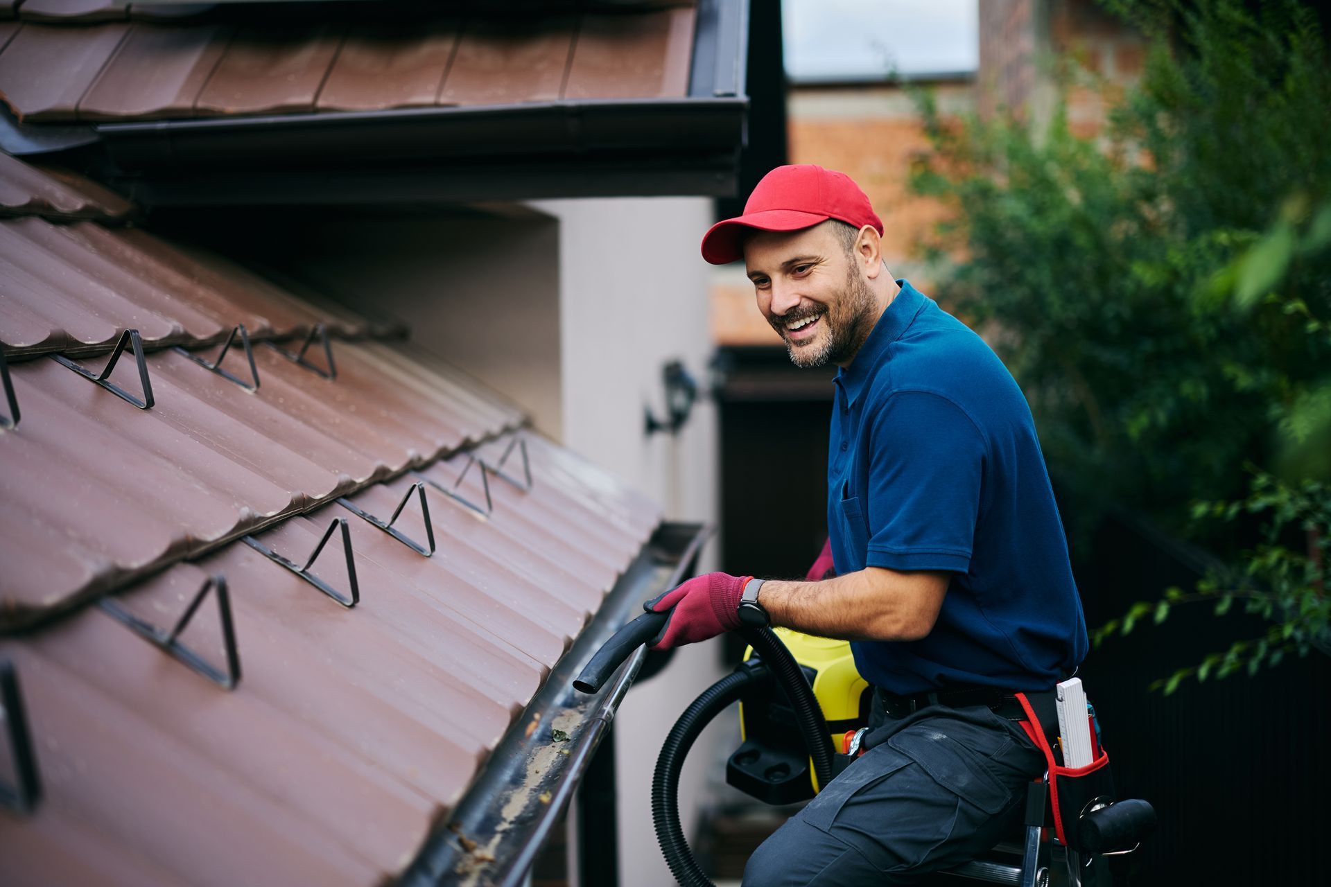 A smiling technician in a red cap and gloves is servicing gutters by a metal roof. A smiling technician in a red cap and gloves is servicing gutters by a metal roof.