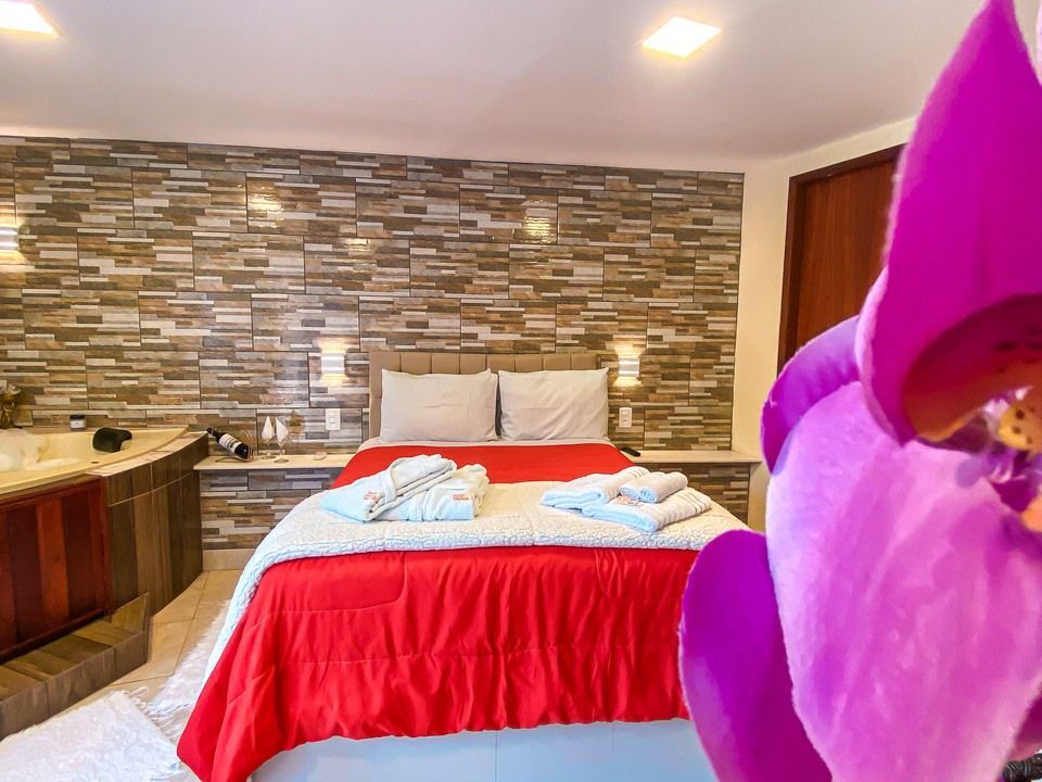 Um quarto com uma cama coberta de vermelho, parede de pedra empilhada e uma banheira de hidromassagem, parcialmente escondida por uma pétala de flor roxa.