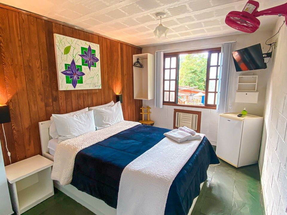 Um quarto de hotel aconchegante com paredes revestidas de madeira, uma cama de casal com roupa de cama azul, uma janela, um frigobar e um ventilador de teto.