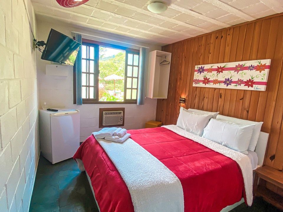 Um quarto de hotel com cama queen-size, edredom vermelho, parede com painéis de madeira, televisão e janela com vista para o jardim.