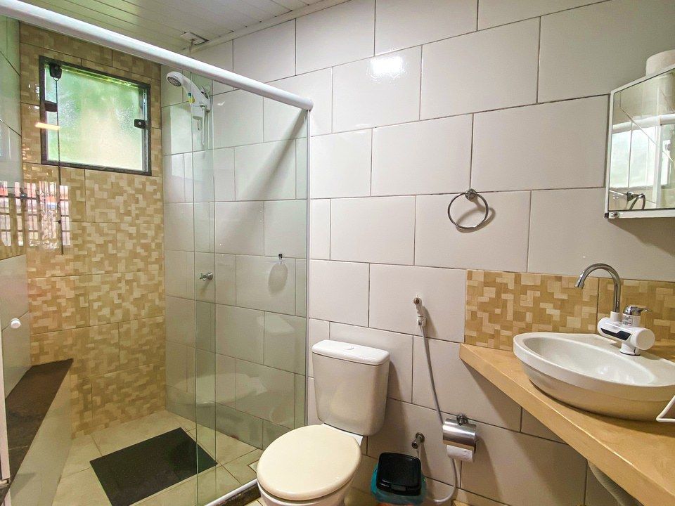 Um banheiro com box de vidro, vaso sanitário branco, pia moderna sobre bancada de madeira e paredes revestidas com azulejos em tons neutros.
