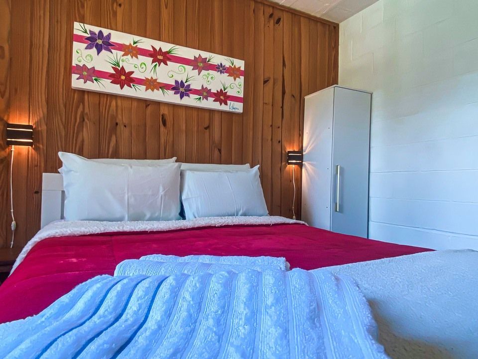 Uma cama arrumada com uma colcha vermelha e um cobertor azul texturizado, encostada em uma parede com painéis de madeira, decorada com arte floral e luminárias de parede.