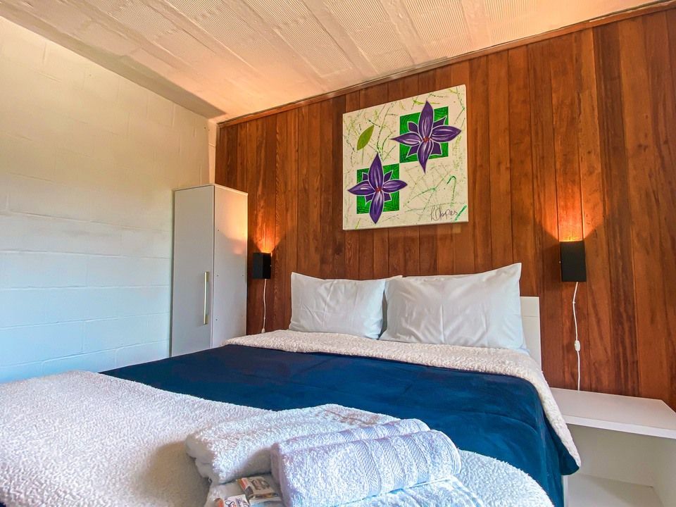 O quarto possui uma cama queen-size com um cobertor azul-marinho, travesseiros brancos, toalhas brancas dobradas e uma obra de arte floral.