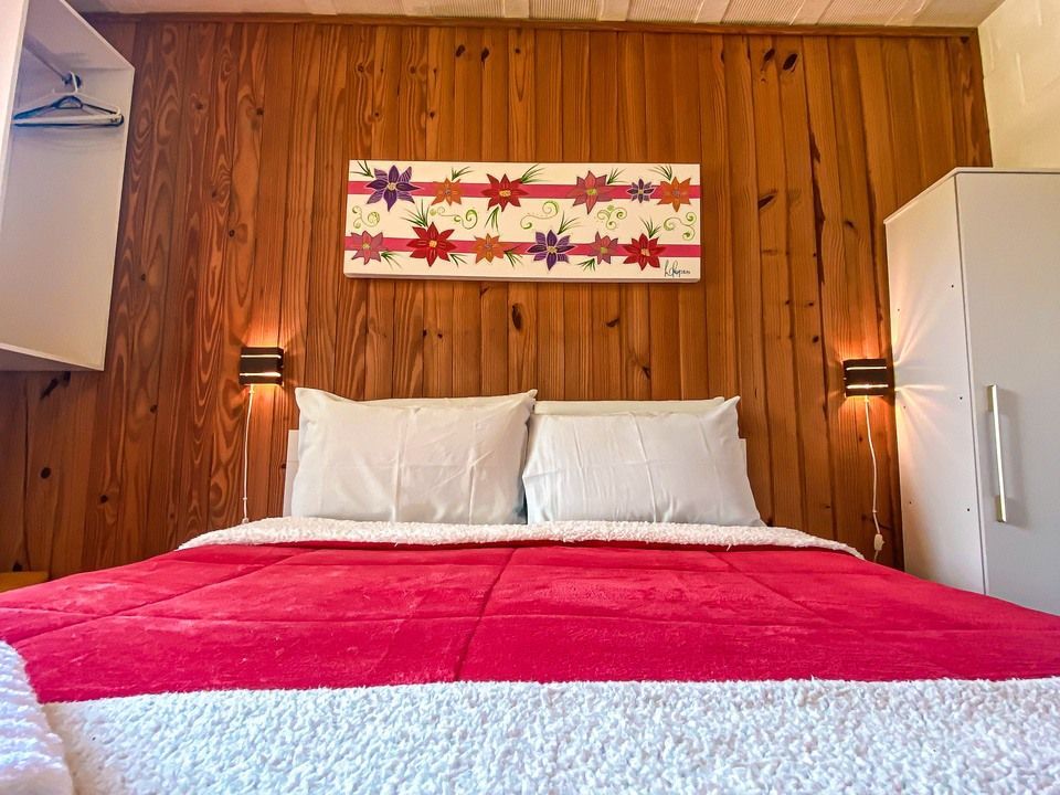 Um quarto com uma colcha vermelha vibrante, dois travesseiros brancos, paredes com painéis de madeira e uma pintura floral acima da cama.