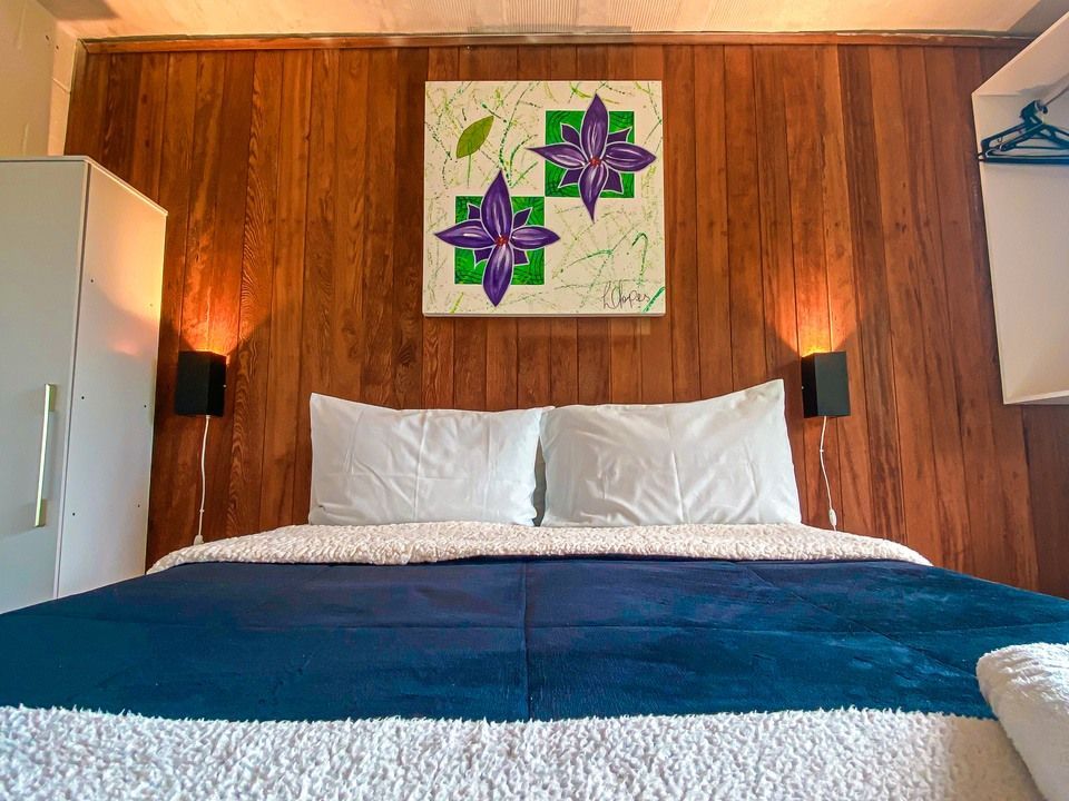 Uma cama com lençóis azuis e travesseiros brancos está encostada em uma parede com painéis de madeira, decorada com uma pintura floral e dois abajures.