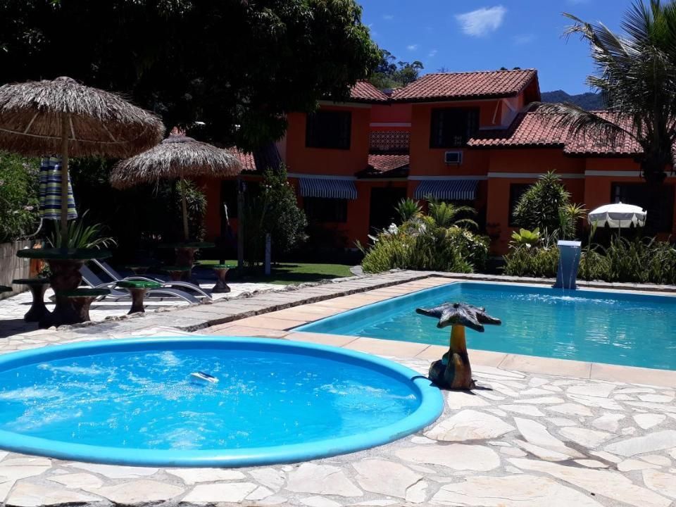 Uma banheira de hidromassagem circular e uma piscina retangular em um pátio com guarda-sóis de palha e uma casa laranja ao fundo.