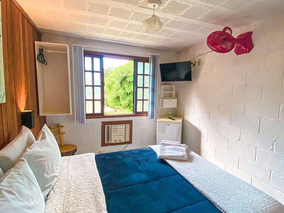 Um quarto de hotel mobiliado dispõe de uma cama queen-size com colcha azul, janela com moldura de madeira, frigobar e TV de parede.