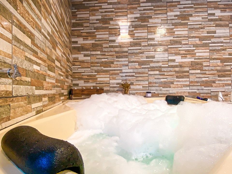 Uma banheira cheia de espuma com encosto de cabeça preto contra uma parede revestida com azulejos texturizados que imitam pedra em várias cores.
