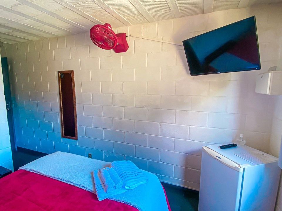 Um quarto de hotel com uma cama coberta com um lençol vermelho, uma parede de blocos de concreto brancos, um pequeno ventilador de parede vermelho, uma TV e um frigobar.