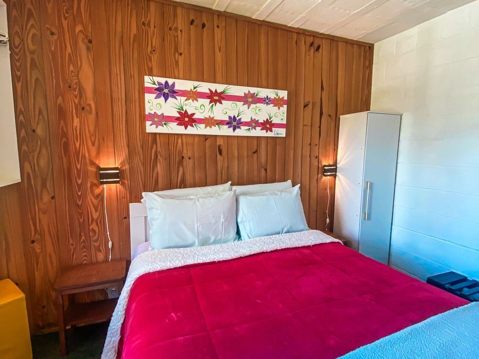 Um quarto com uma cama coberta por um cobertor vermelho vibrante, travesseiros azul-claros, paredes com painéis de madeira e uma pintura floral na parede.