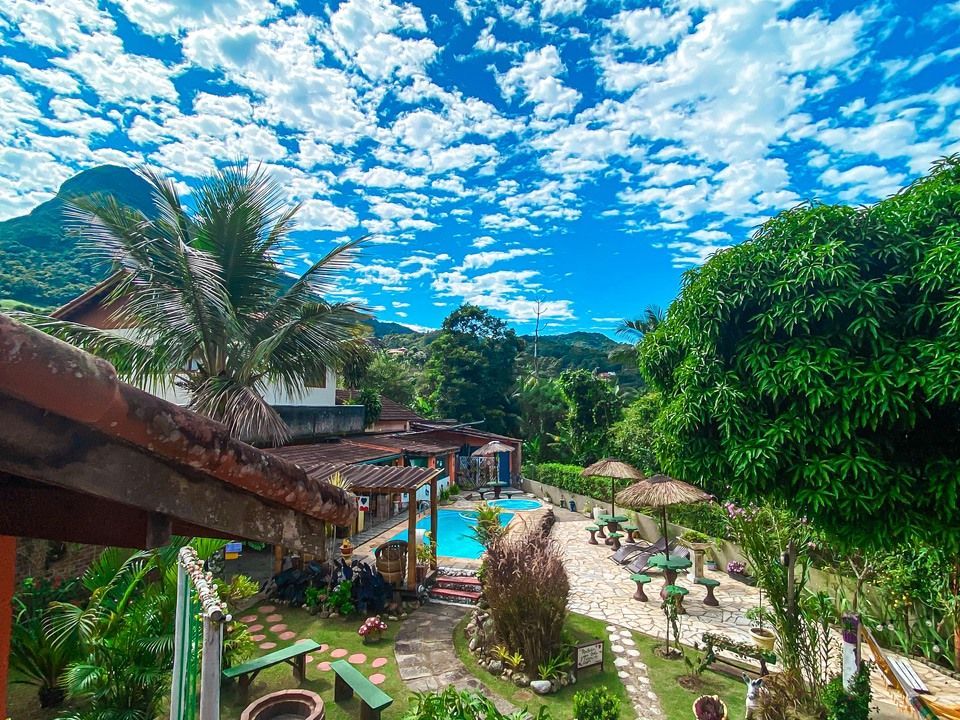 Um resort tropical com piscina rodeada por vegetação exuberante, caminhos de pedra e montanhas sob um céu azul.