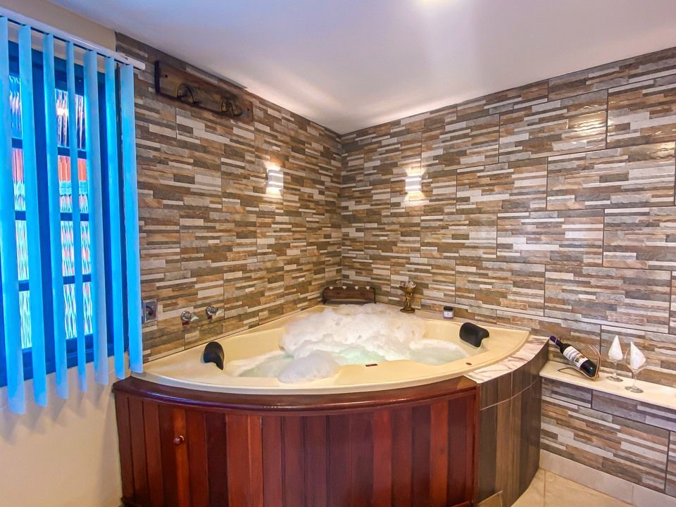 Uma banheira de canto com hidromassagem e espuma de banho, ao lado de uma janela com persianas verticais azuis e paredes revestidas com azulejos de pedra.