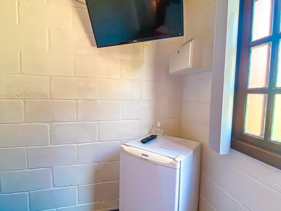 Uma televisão fixada na parede está suspensa acima de um pequeno frigobar branco em uma sala com paredes de tijolos pintados de branco e uma janela.