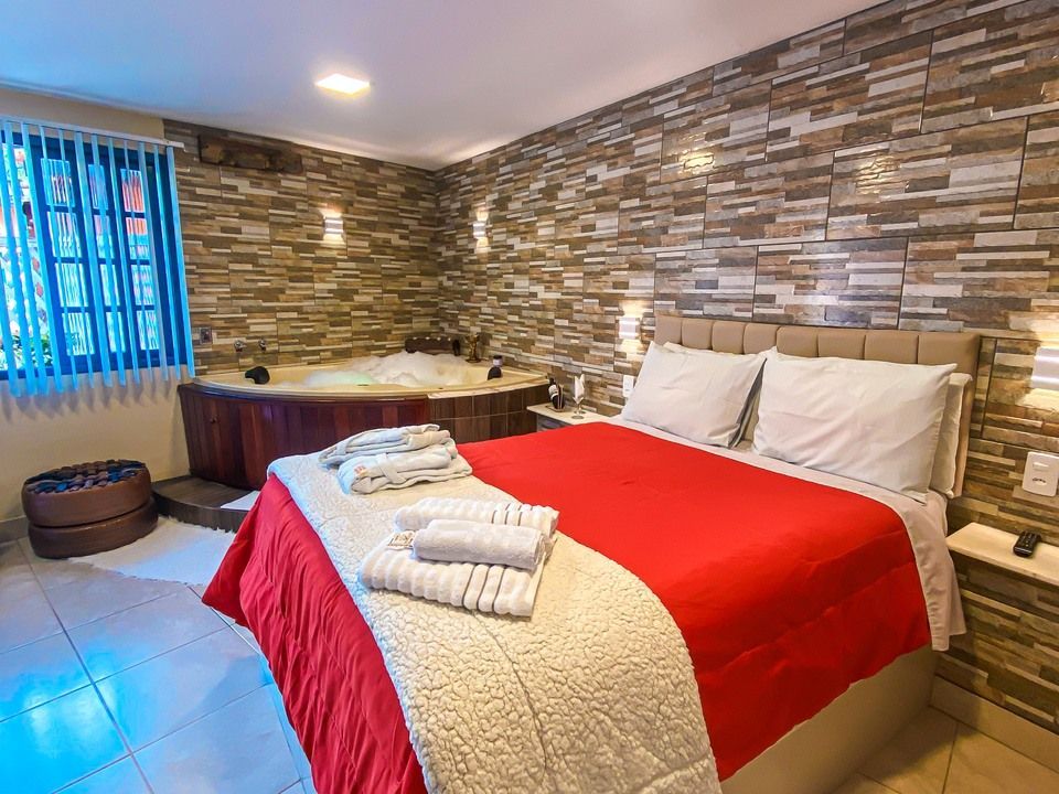 Um quarto de hotel com uma cama com um edredom vermelho vivo e uma jacuzzi embutida contra uma parede de pedra texturizada.