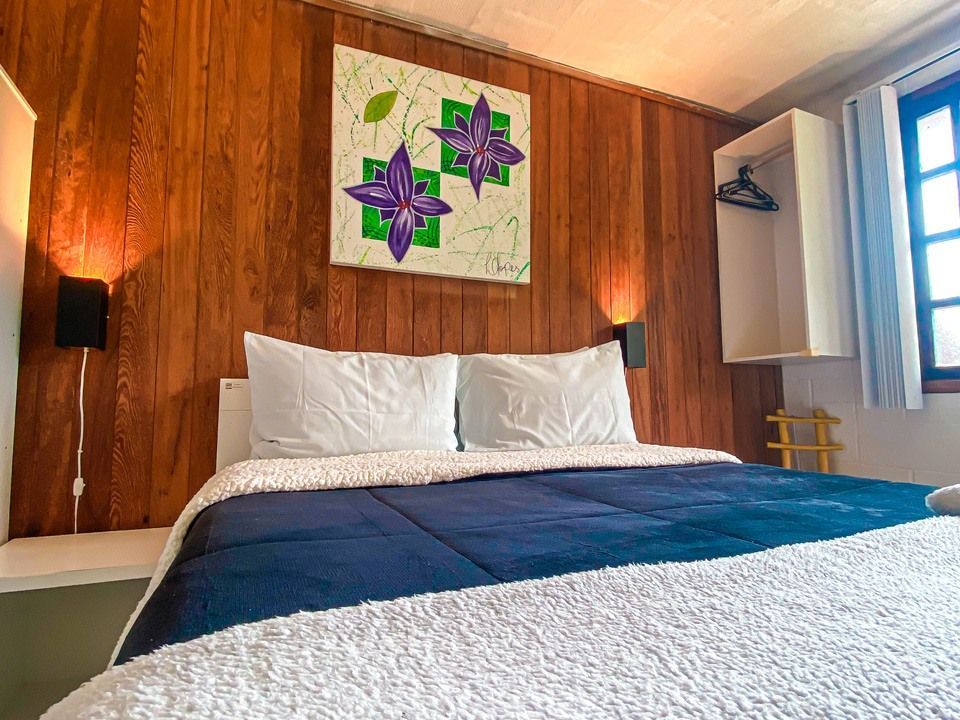 Uma cama com travesseiros brancos e um edredom azul encostada em uma parede de madeira com uma obra de arte floral.