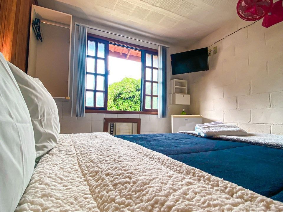 Um quarto luminoso com uma cama com um cobertor branco texturizado e roupa de cama azul-escura, uma janela com vegetação e uma TV.