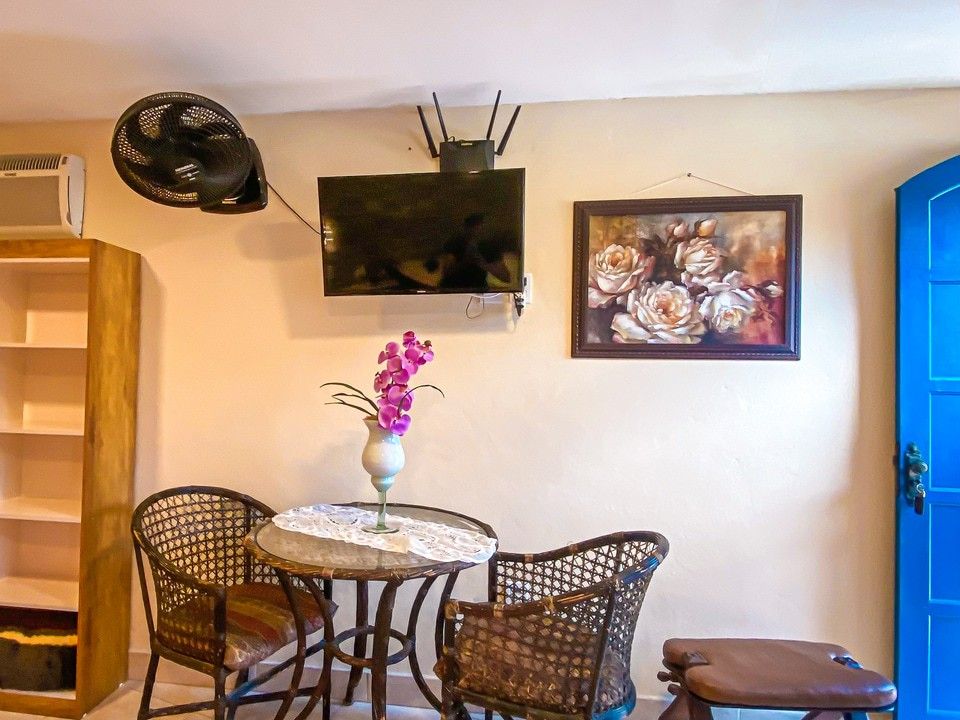 Uma sala com uma pequena mesa de bistrô e duas cadeiras de vime, uma TV fixada na parede, um vaso de flores e um quadro na parede.