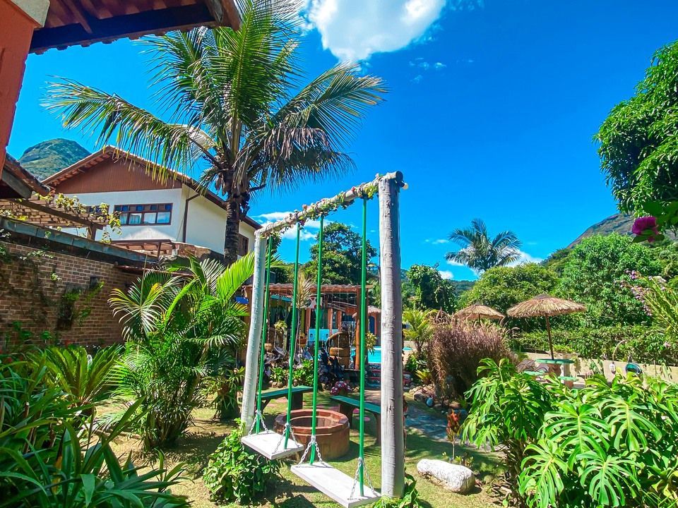 Um jardim ensolarado e exuberante com um balanço de madeira, plantas tropicais, árvores e uma casa sob um céu azul límpido.