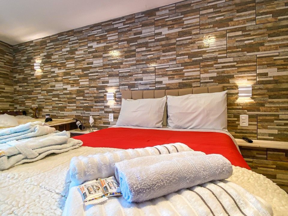 Uma cama de hotel com colcha vermelha e lençóis brancos, com uma parede de destaque em padrão de pedra e apliques de parede modernos.