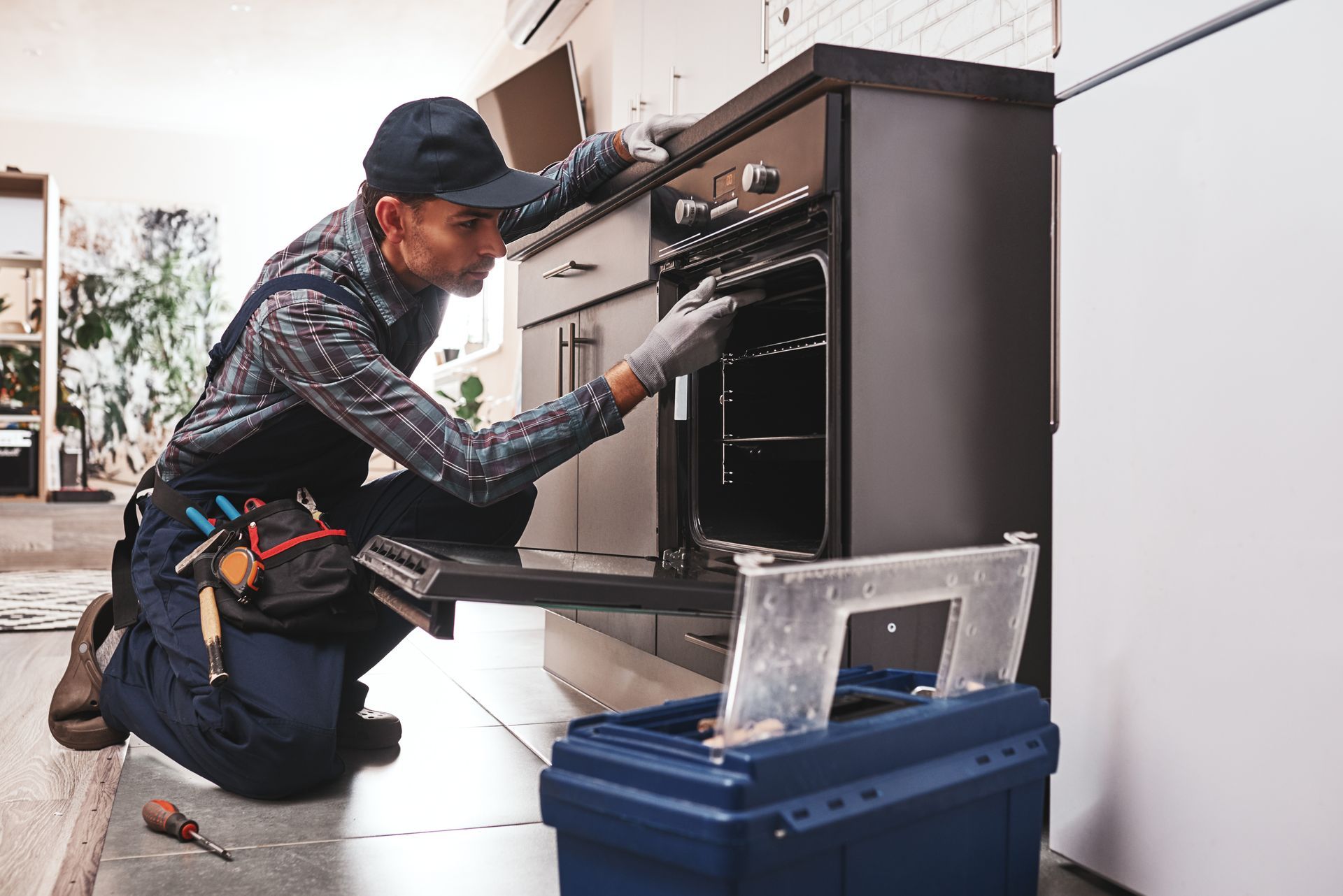 A man is kneeling down in a kitchen fixing an oven.