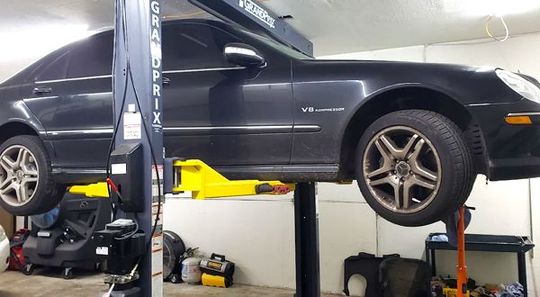 Black Mercedes sedan on a lift in a garage. Yellow lift arms, tools, and equipment are visible.