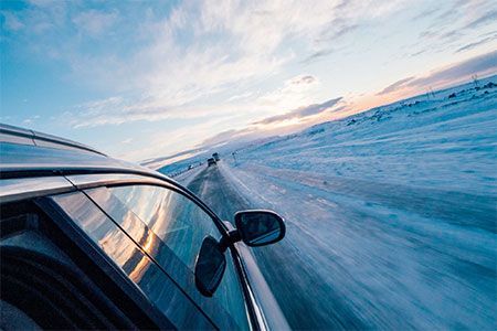 Car driving on a snow-covered road under a blue sky, with mountains in the distance.