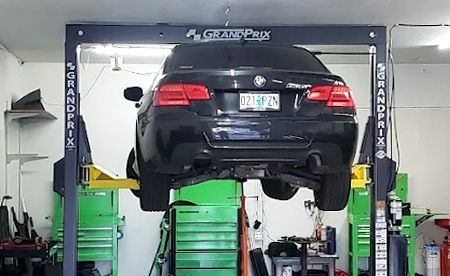 Black car raised on a Grand Prix car lift in a garage with green tool chests.