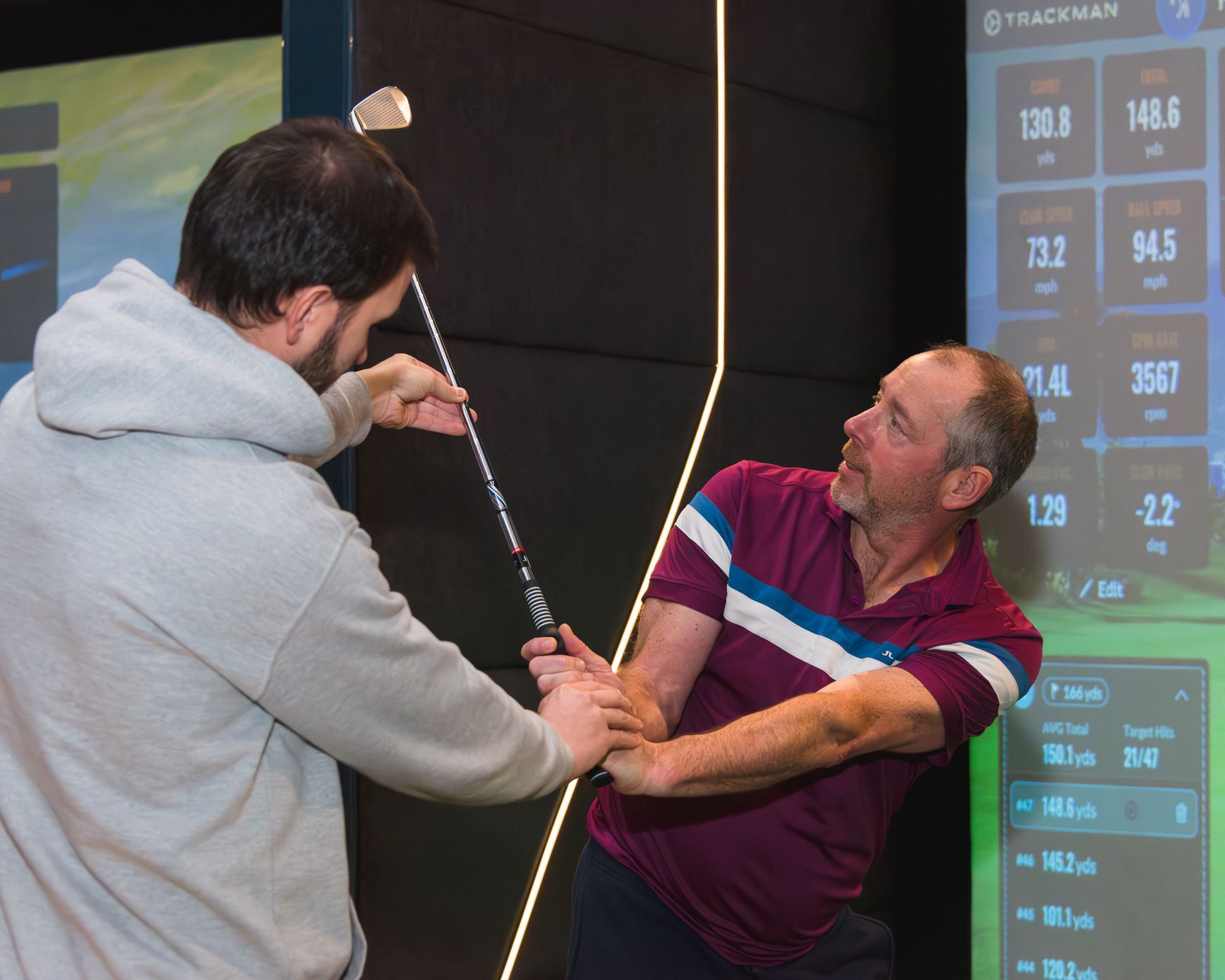 Woman swinging golf club indoors, observed by two people, green screen golf course in background.