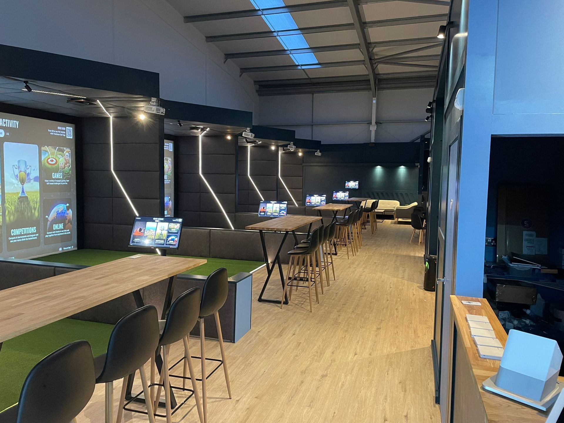 Interior of a golf simulator facility with bays, screens, seating, and a bar area.