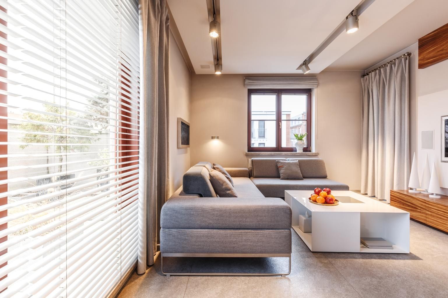 Modern living room with gray sofa, white coffee table, and large windows with blinds