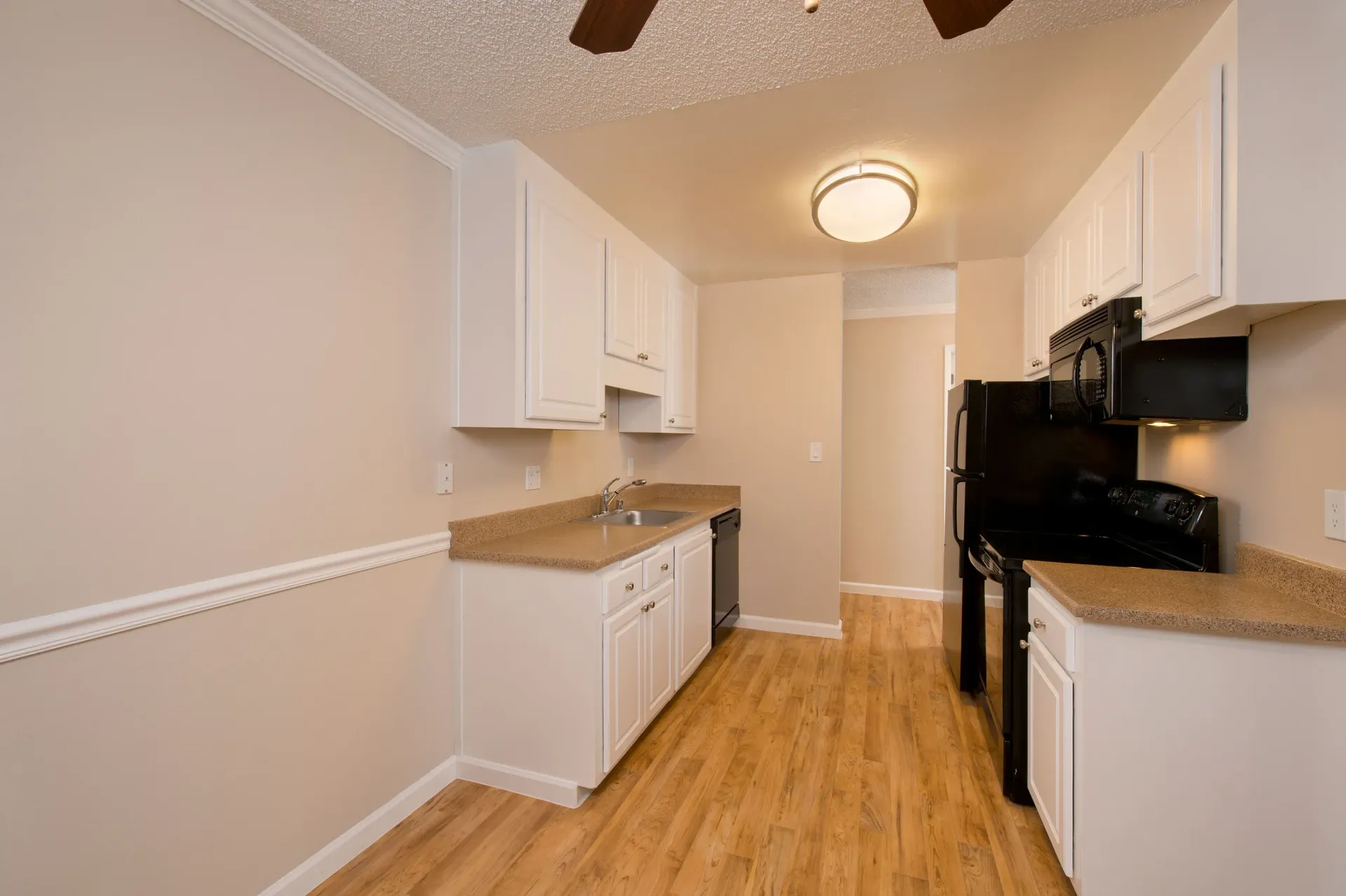 Galley-style apartment kitchen with white cabinets, beige countertops, wood flooring, and black appliances.