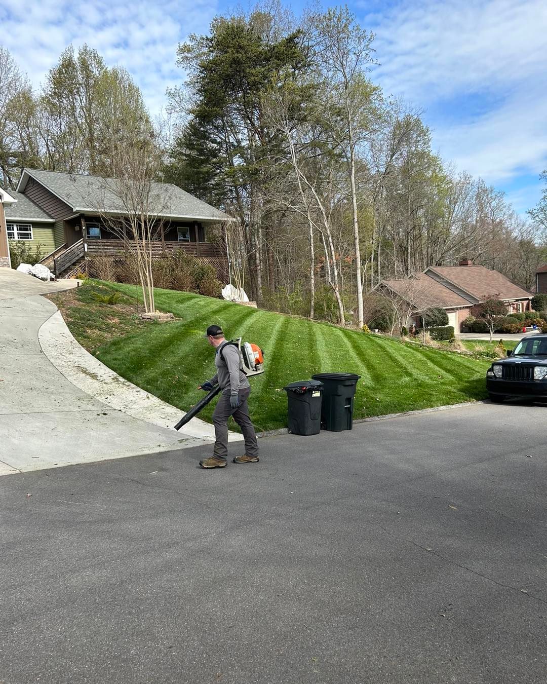 Man using a leaf blower on a driveway next to a neatly mowed lawn with a house in the background.
