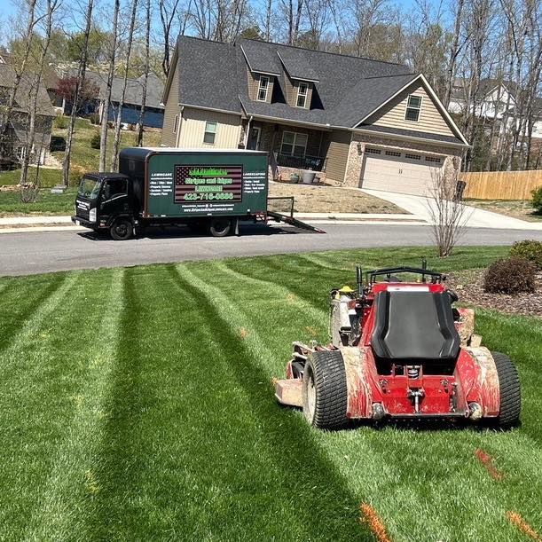 A red lawnmower mowing a striped green lawn near a black truck in front of a house.