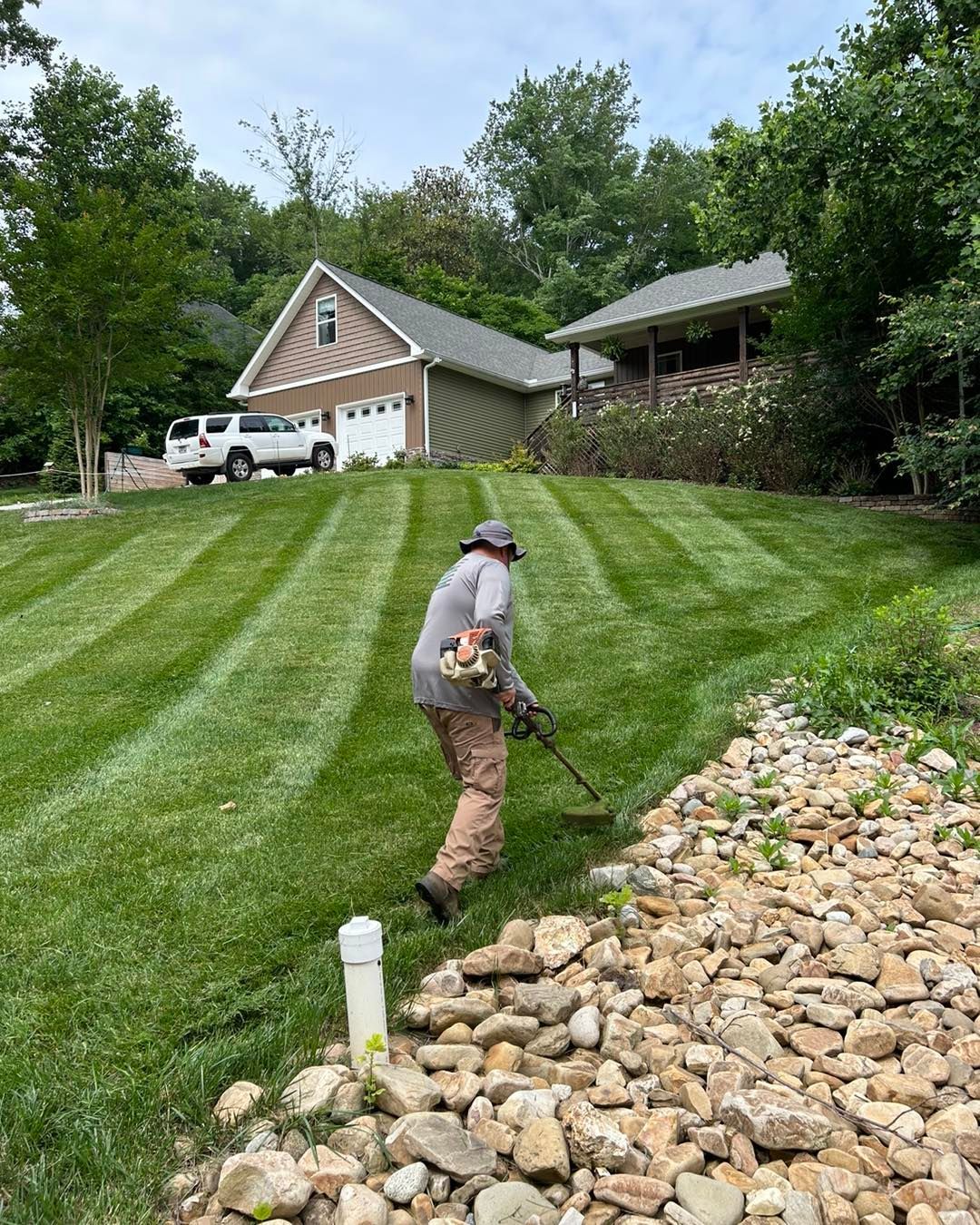Person using a weed eater to trim grass near a rocky drainage area on a sloped lawn with striped patterns in front of a house.