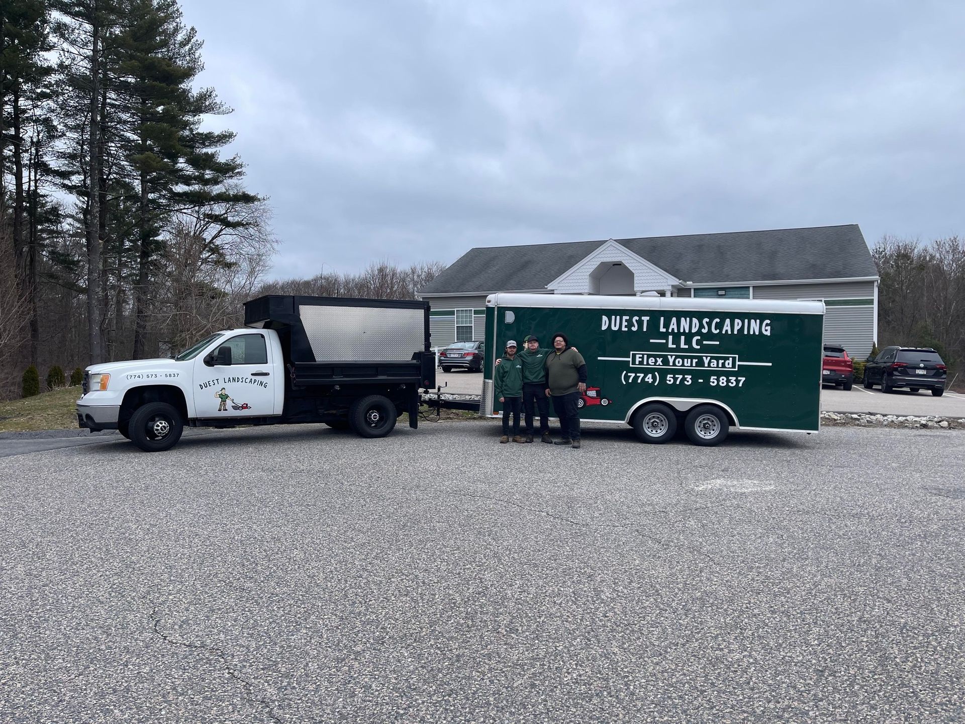 Three people stand by a landscaping truck and trailer in front of a building. Cloudy sky, gravel parking lot.
