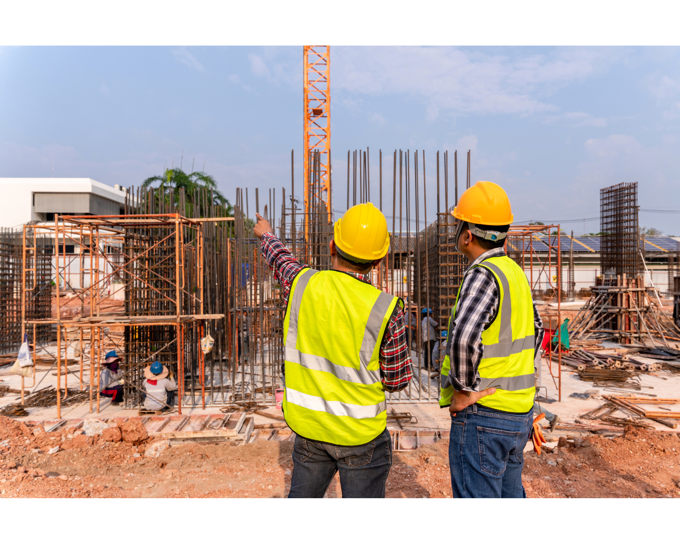 Two construction workers are standing at a construction site looking at a crane.