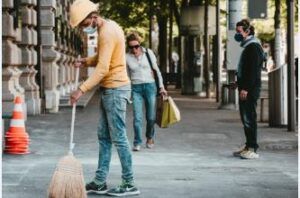 A man wearing a mask is sweeping the sidewalk with a broom.