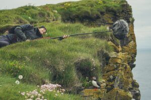 A man is laying on top of a grassy hill next to a cliff.