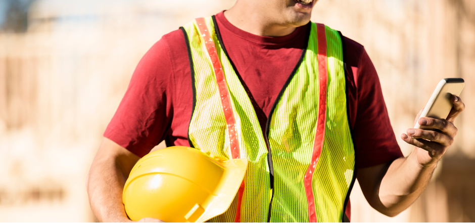 A construction worker is looking at his cell phone at a construction site.